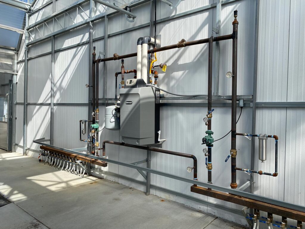 A photo of a greenhouse heating system with pipes and a main unit attached to an outside wall.